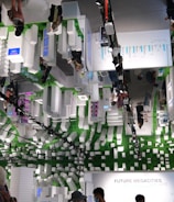 An indoor exhibition space with a futuristic theme featuring white, block-like structures and several people moving through the area. The ceiling mirrors the scene below, adding to the visual complexity. Green elements and digital display panels are integrated into the design. The space has an informational focus, likely about urban development or technology.