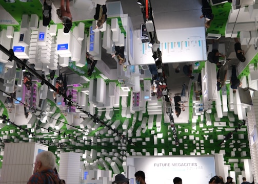 An indoor exhibition space with a futuristic theme featuring white, block-like structures and several people moving through the area. The ceiling mirrors the scene below, adding to the visual complexity. Green elements and digital display panels are integrated into the design. The space has an informational focus, likely about urban development or technology.