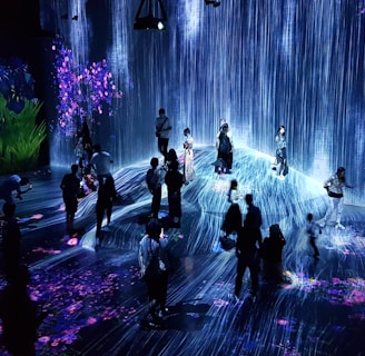 A group of people are standing and walking inside an immersive art installation with flowing digital streams and vibrant floral projections on the walls and floor. The environment appears interactive, with the participants seemingly engaged with the digital elements around them.
