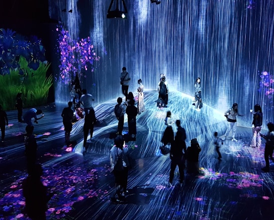 A group of people are standing and walking inside an immersive art installation with flowing digital streams and vibrant floral projections on the walls and floor. The environment appears interactive, with the participants seemingly engaged with the digital elements around them.