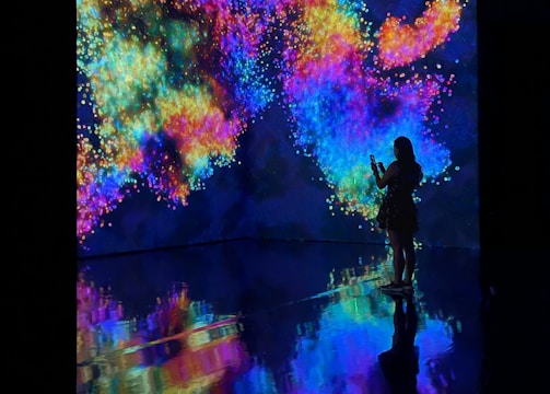 An immersive digital art installation features vibrant, swirling colors projected onto walls. A person stands in silhouette against this vivid backdrop, taking a photo or video. The floor reflects the colorful patterns, creating a mirrored effect.