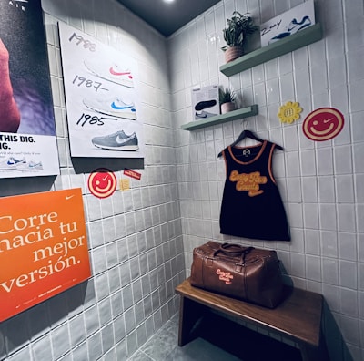 A small retro-themed display with a focus on athletic apparel and shoes. The walls feature a series of posters showcasing various Nike shoes from different years. A black gym tank top and a brown leather gym bag are prominently positioned on a wooden bench. There are decorative elements like potted plants on wall-mounted shelves and smiley face icons. The environment has a nostalgic and sporty atmosphere.