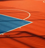 A vibrant sports court with sections of red and blue, featuring crisp white lines marking the boundaries. Shadows from a structure create geometric patterns across the surface, adding depth and interest to the scene.