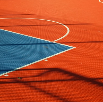 A vibrant sports court with sections of red and blue, featuring crisp white lines marking the boundaries. Shadows from a structure create geometric patterns across the surface, adding depth and interest to the scene.
