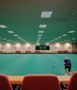 A large indoor sports facility featuring a green playing surface, similar to a bowling green. The ceiling is lined with numerous fluorescent lights, giving the space a bright yet serene atmosphere. Red chairs are arranged along the walls, and a scoreboard can be seen in the distance. A person is actively engaged in the game, bending forward as they release a ball, while inset a few spectators or participants are visible on the left side of the space.