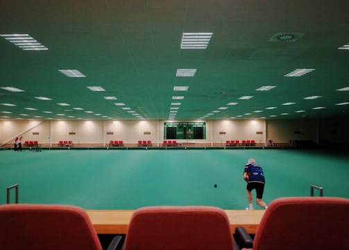 A large indoor sports facility featuring a green playing surface, similar to a bowling green. The ceiling is lined with numerous fluorescent lights, giving the space a bright yet serene atmosphere. Red chairs are arranged along the walls, and a scoreboard can be seen in the distance. A person is actively engaged in the game, bending forward as they release a ball, while inset a few spectators or participants are visible on the left side of the space.